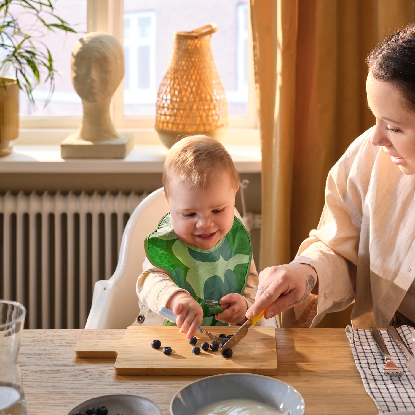 A toddler wearing a GRÖNFINK bib sits at a dining table holding SMASKA cutlery, picking up soft fruit from a chopping board.