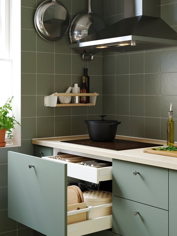 Cuisine carrelée de vert et équipée d’armoires avec façades NICKEBO gris-vert, d’une hotte et d’une table de cuisson noire sur laquelle est posée une cocotte en fonte.