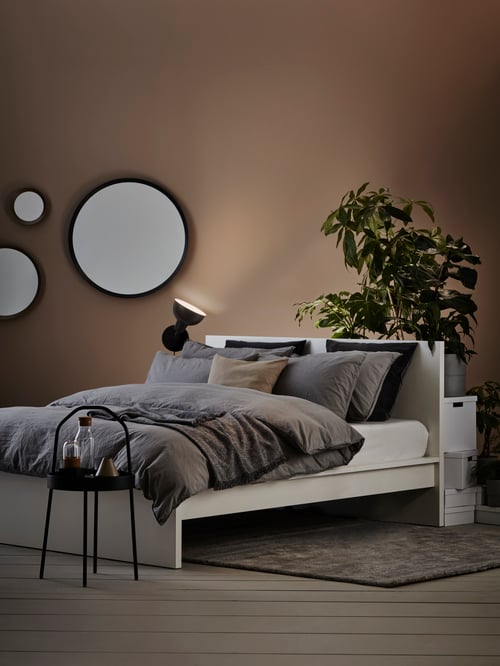 Bedroom with bed frame, duvet cover, pillowcases, side table, mirrors, cushion covers, rug, boxes, plants, wall uplighter.