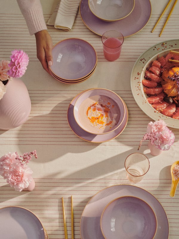 A person puts a stack of bowls, part of a light pink FÄRGKLAR 18-piece service set, by a vase while setting a table.