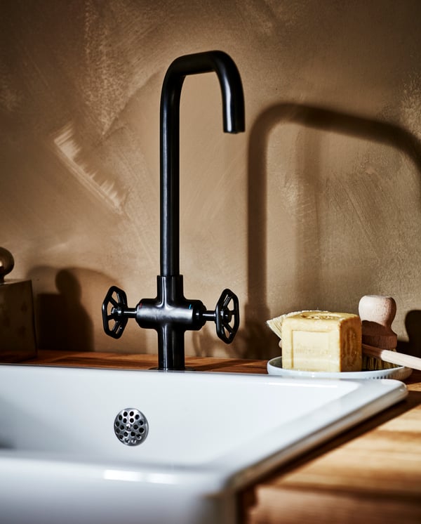 A dual-control kitchen mixer tap in black brushed metal, a white sink bowl, a worktop in oak/veneer and a dish brush in wood.