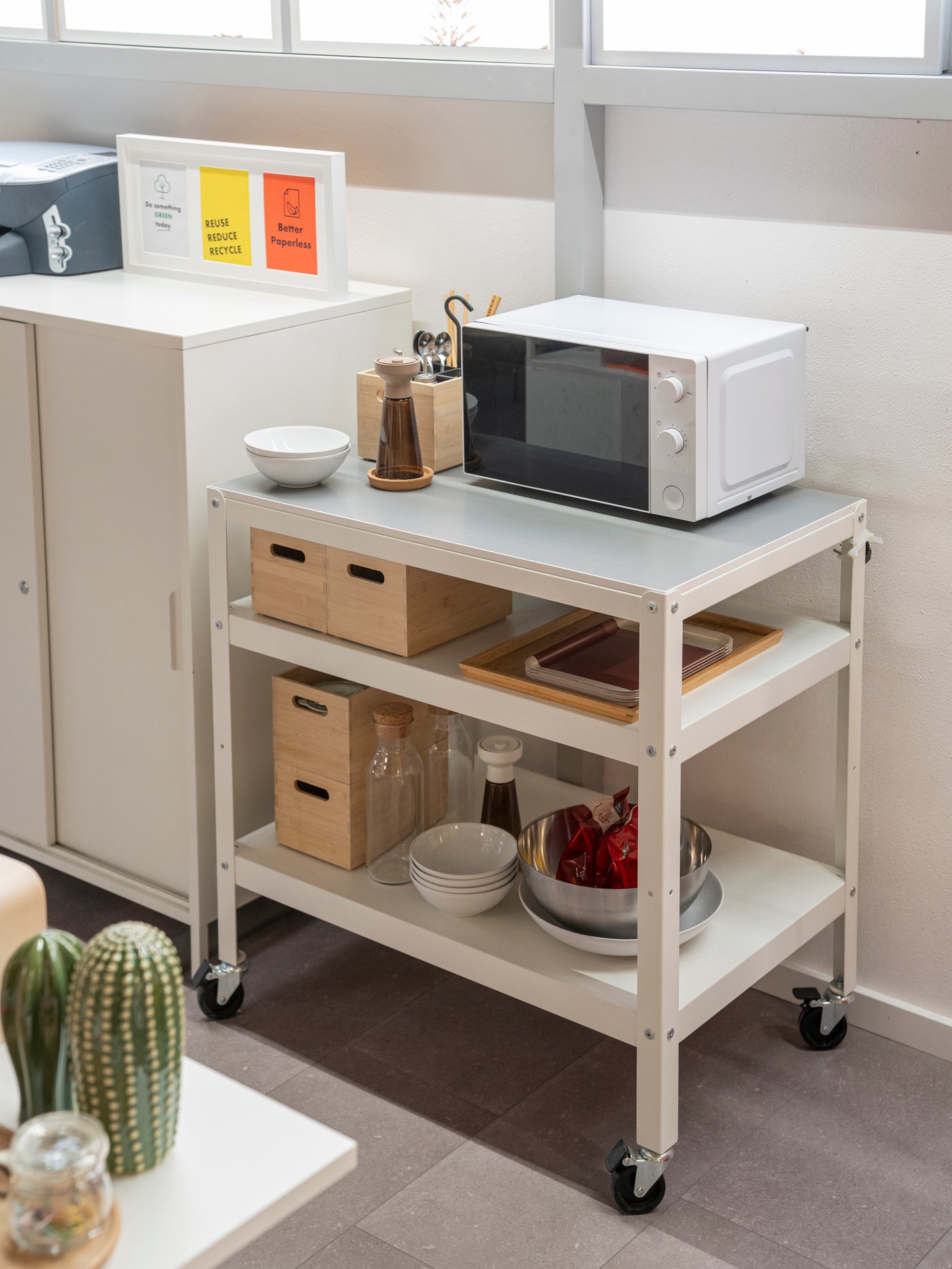 Vit serveringsvagn på hjul med mikrovågsugn, förvaringslådor och köksartiklar i ett ljust pentry eller kontor.