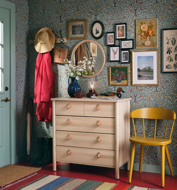 En ljus träbyrå med läderhandtag står i en hall till höger om ytterdörren, dekorerad med blå vas med vita blommor, lampa och småsaker. Ovanför hänger en rund spegel och flera inramade bilder. Till vänster står en klädhängare med hattar och jackor, och till höger en gul stol. Väggarna har en blommig tapet och randiga mattor ligger på golvet.