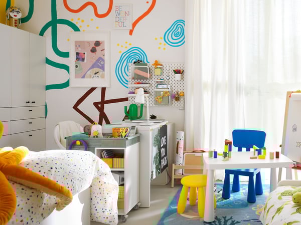 Children’s room with big colourful drawings on the wall behind a desk, a small play table, toys and craft everywhere.