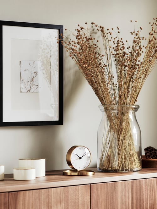 Une photo encadrée au-dessus d’une surface en bois sur laquelle se trouvent des boîtes de décoration, un réveil et des branches artificielles dans un vase.