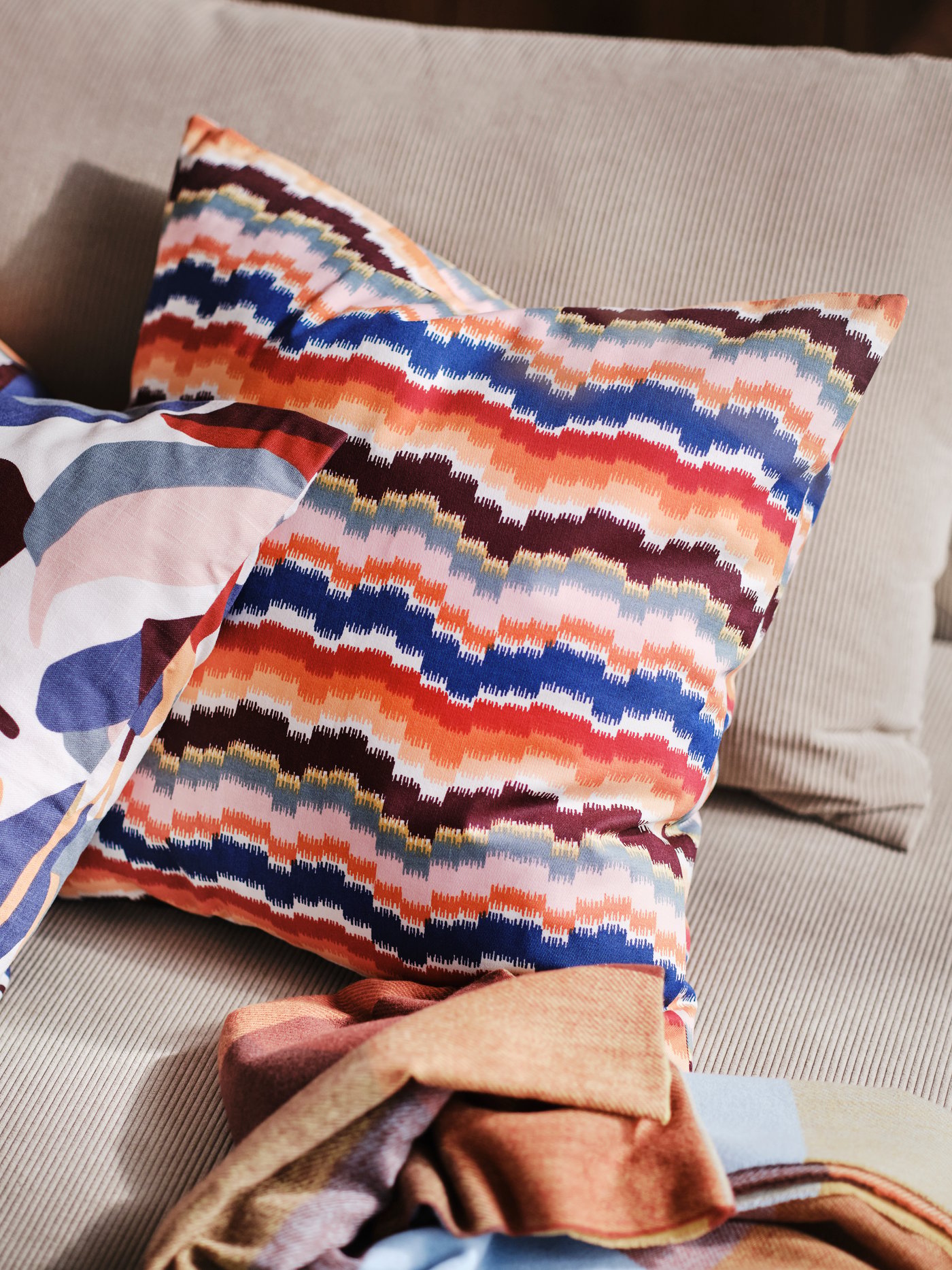 A close-up of two cushions on a beige sofa. One has a multicoloured PEKINGSYREN cushion cover with a zigzag pattern.