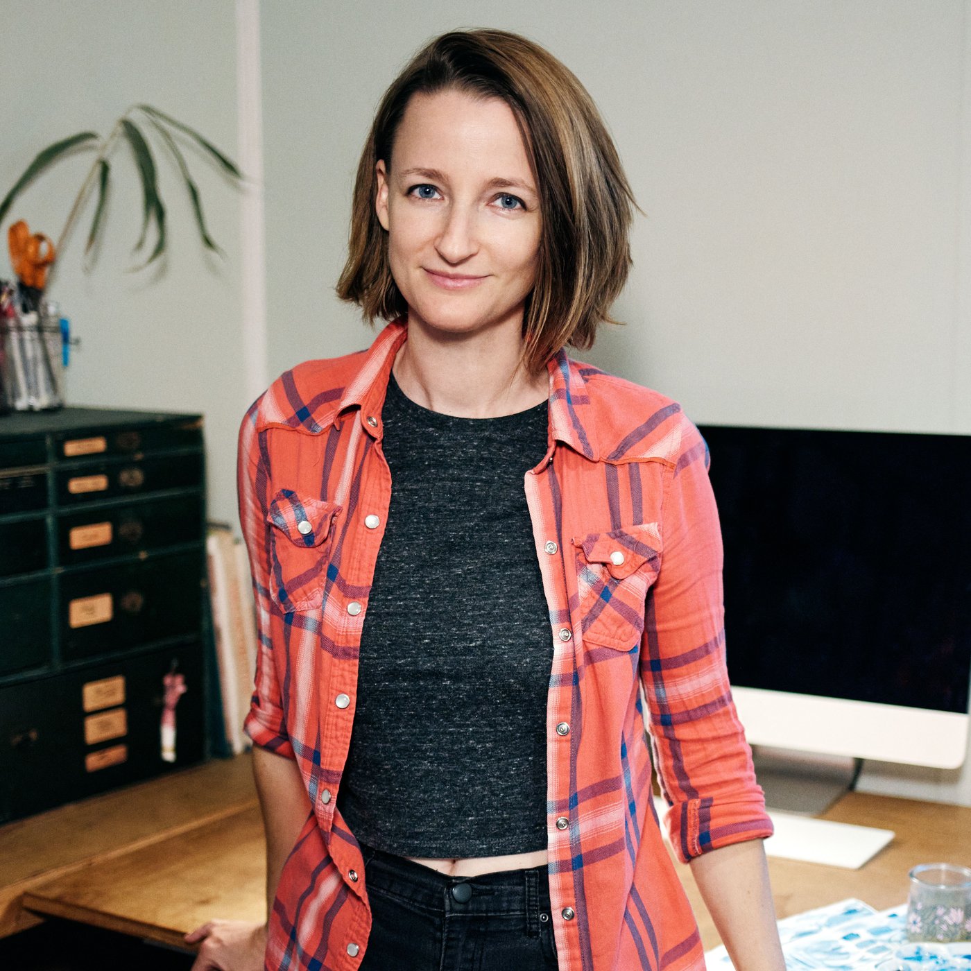 A woman standing in front of a computer on a desk full of pencils in pencil stands. To the left is a medium shelving unit with storage boxes on top.