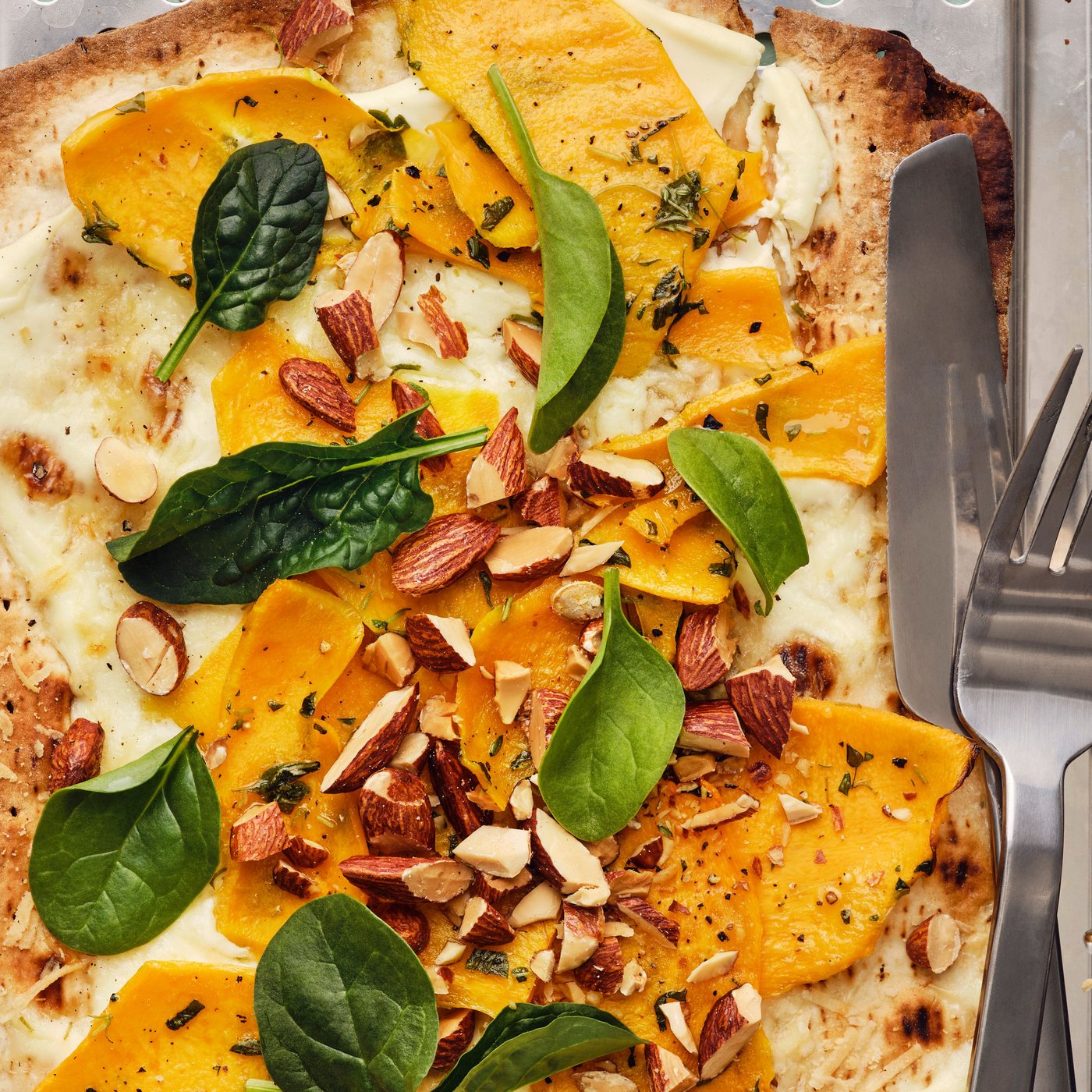 Pizza with GRÄDDAT soft thin flatbread and roasted pumpkin, topped with spinach, and MUNSBIT roasted almonds, with cutlery.