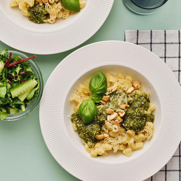 To hvite dype tallerkener fylt med BÄSTISAR pasta, grønn pesto og hakkede cashewnøtter på en lysegrønn overflate.
