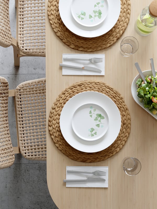 Ein stilvoll gedeckter Tisch mit weißen Tellern, die mit zarten grünen Pflanzenmotiven verziert sind. Dazu passende, runde Untersetzer aus Naturmaterial und klare Gläser. Ein frischer Salat in einer modernen Schüssel rundet das Gesamtbild ab, während die Rattanstühle für eine gemütliche Atmosphäre sorgen.