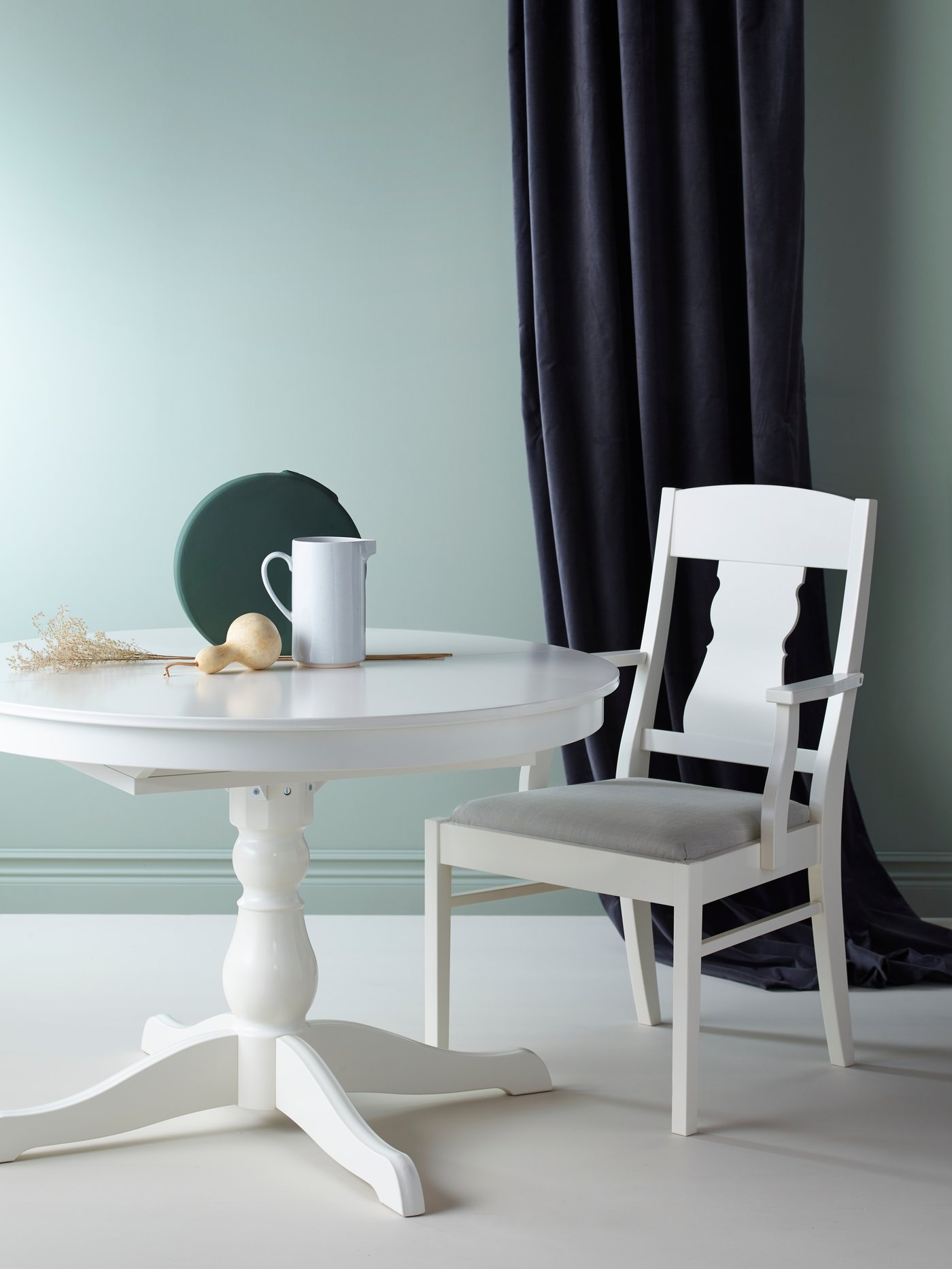 Zona de comedor con una mesa extensible y un jarrón encima, una silla con reposabrazos debajo y un par de cortinas en la parte de atrás.