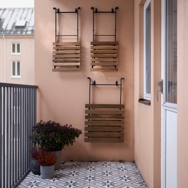 A folded TÄRNÖ table and two folded TÄRNÖ chairs hang on a wall on a balcony near some plants in pots.