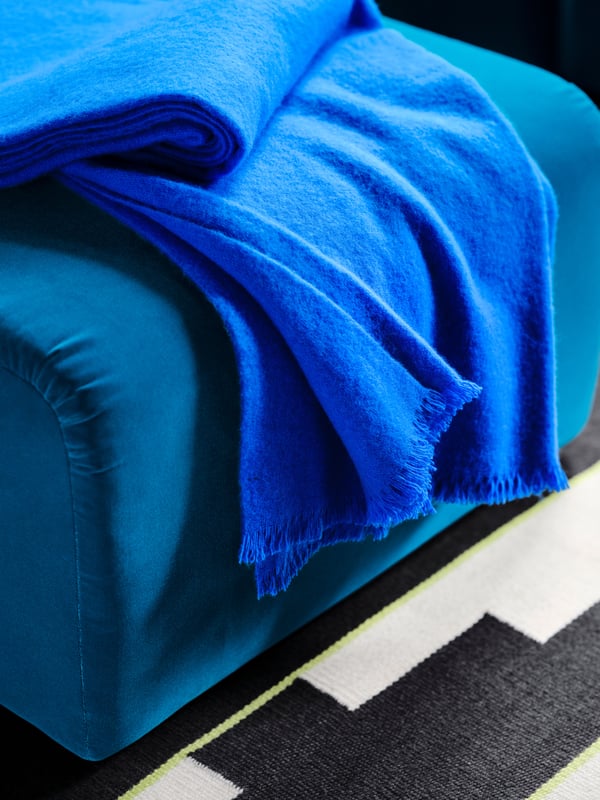 A bright blue STOCKHOLM 2025 throw rests on a dark turquoise pouffe sitting on a black/white and light green handwoven rug.