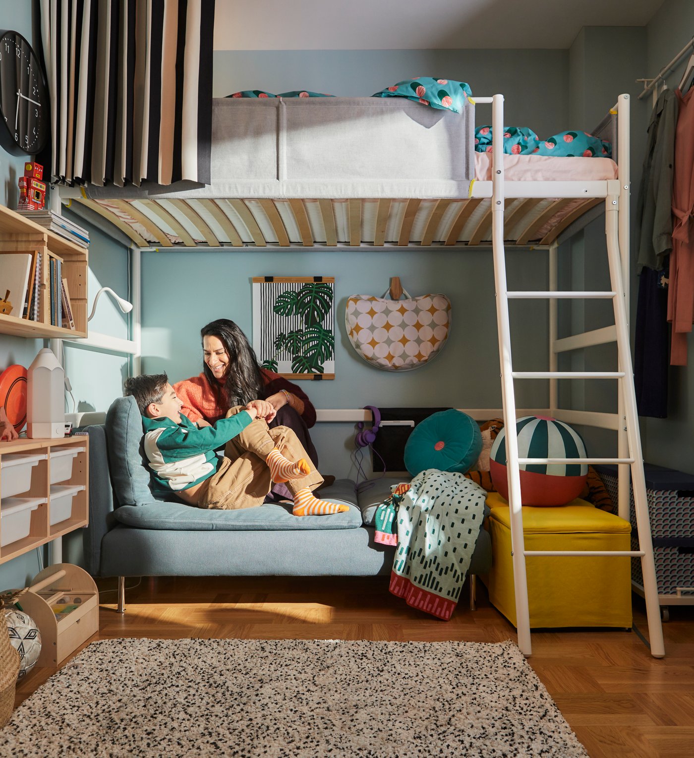VITVAL loft bed frame with a mother and son sitting down on a mini blue couch below it. The walls are blue and decorated with shelves and soft toys.