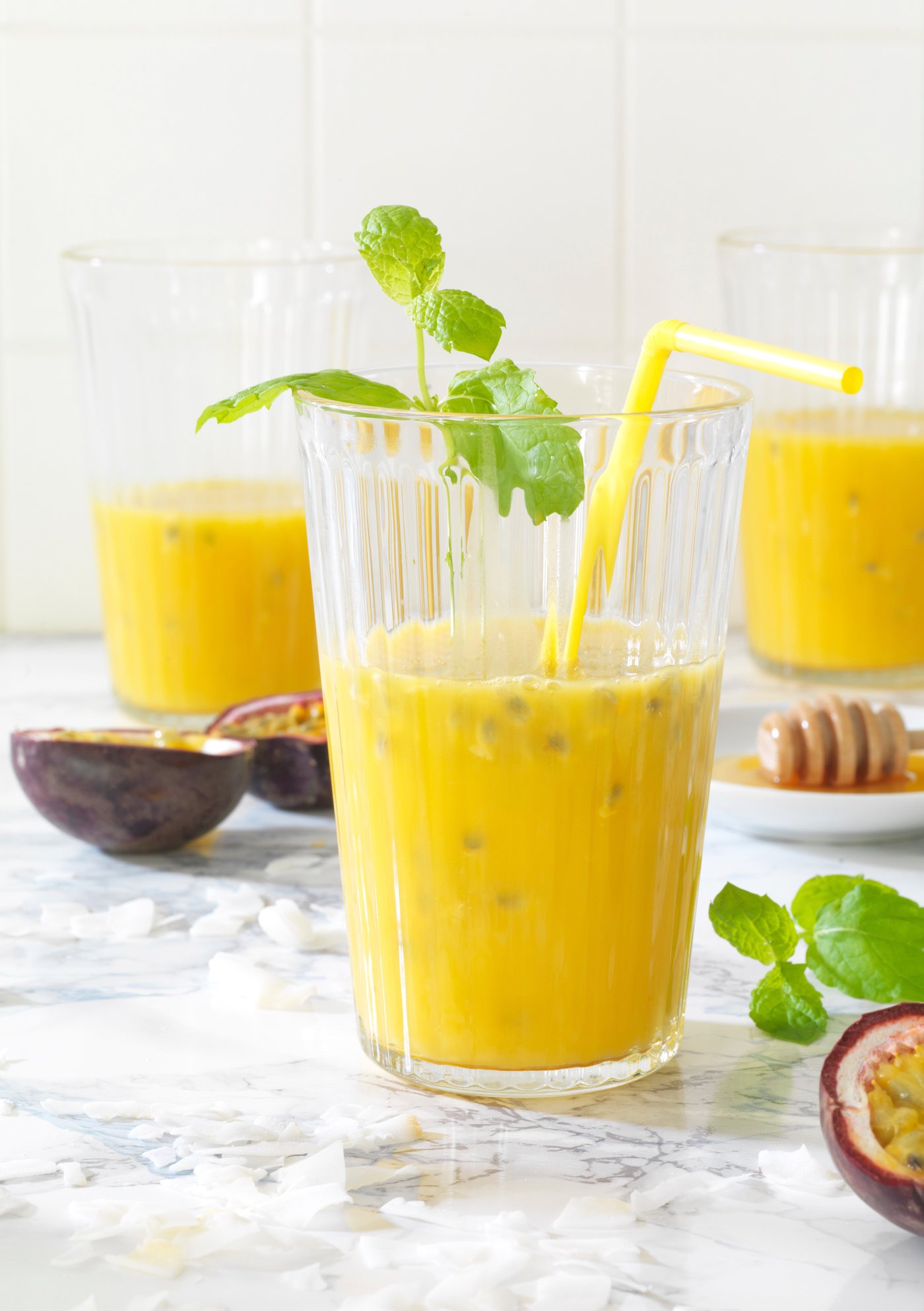 Ein VARDAGEN Glas, gefüllt mit frisch gepresstem Saft, steht auf einer hellen Marmorfläche und ist mit Minzblättern und einem Trinkhalm dekoriert. Im Hintergrund sind weitere Gläser sowie halbierte Passionsfrüchte und Honig zu sehen.