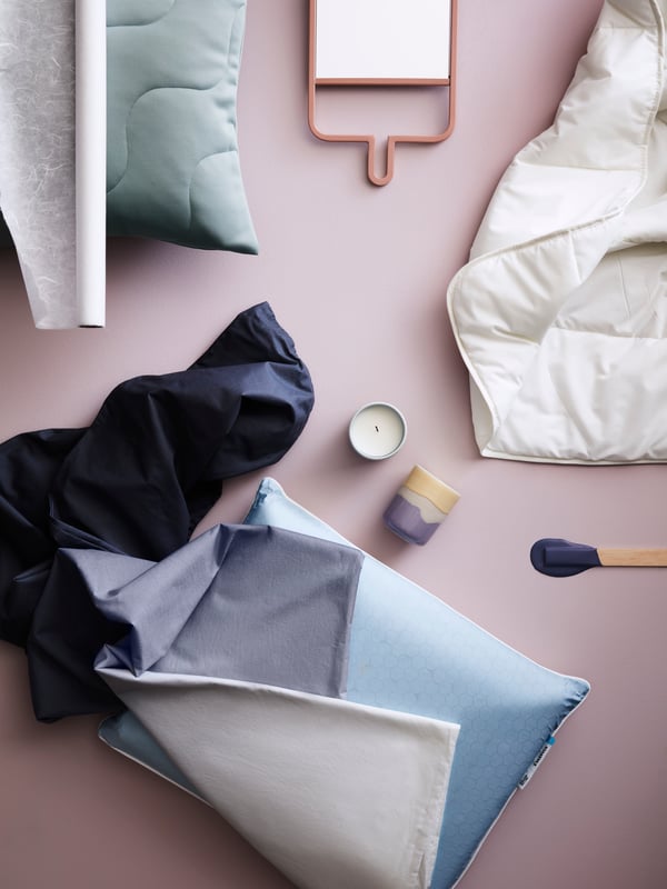 An artistic arrangement of a blue pillow, light-green cushion, white duvet, candle holders and mirror on a pink surface.
