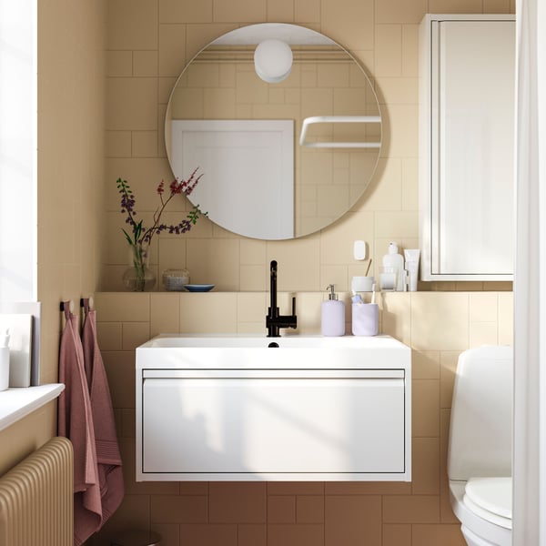 A white ÄNGSJÖN wash-stand with one drawer is mounted in a bathroom with light brown tiles below a round FAXÄLVEN mirror.
