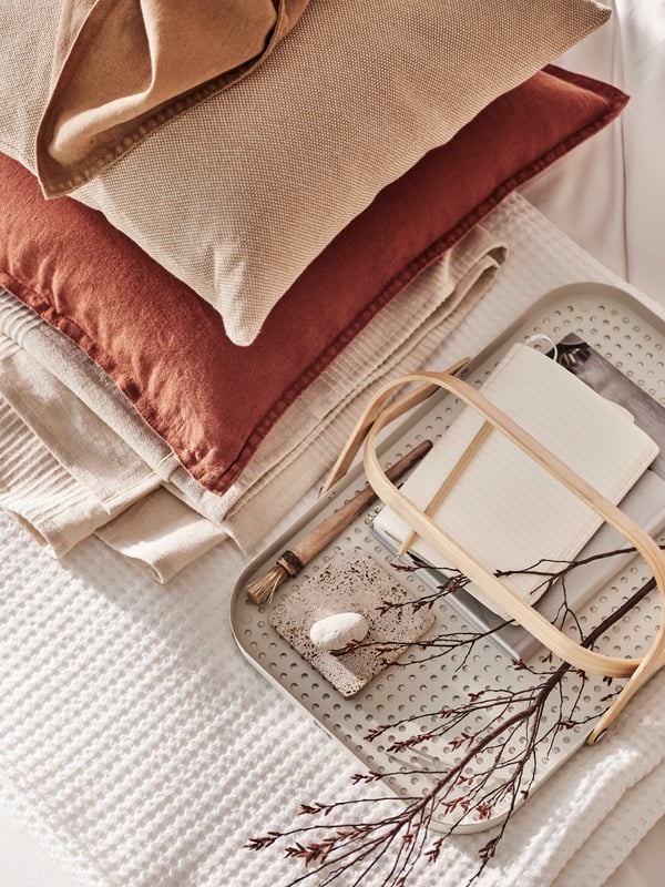 Dous coxíns, un cunha funda DYTÅG en marrón avermellado e outro cunha funda SANDTRAV rosa nunha morea con máis roupa de cama dobrada.