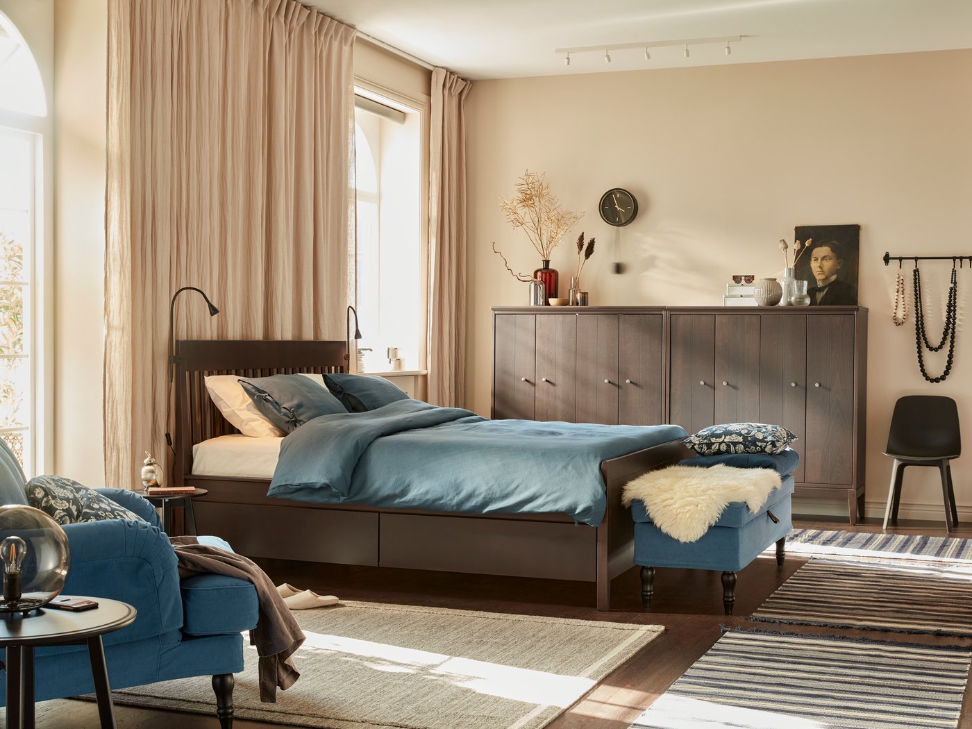 Camera da letto con una struttura letto con contenitori. La stanza ha pareti beige, tende in lino, panca con cuscini, una poltrona, un mobile in legno accostati alla parete e una sedia.