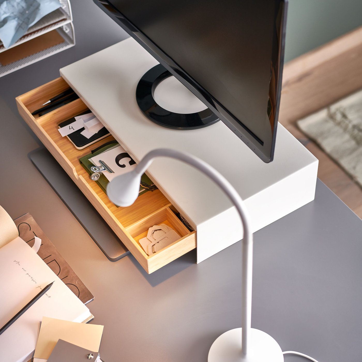 En vit NÄVLINGE LED-arbetslampa står på en grå bordsskiva bredvid ett ELLOVEN bildskärmsstativ med en utdragen låda.