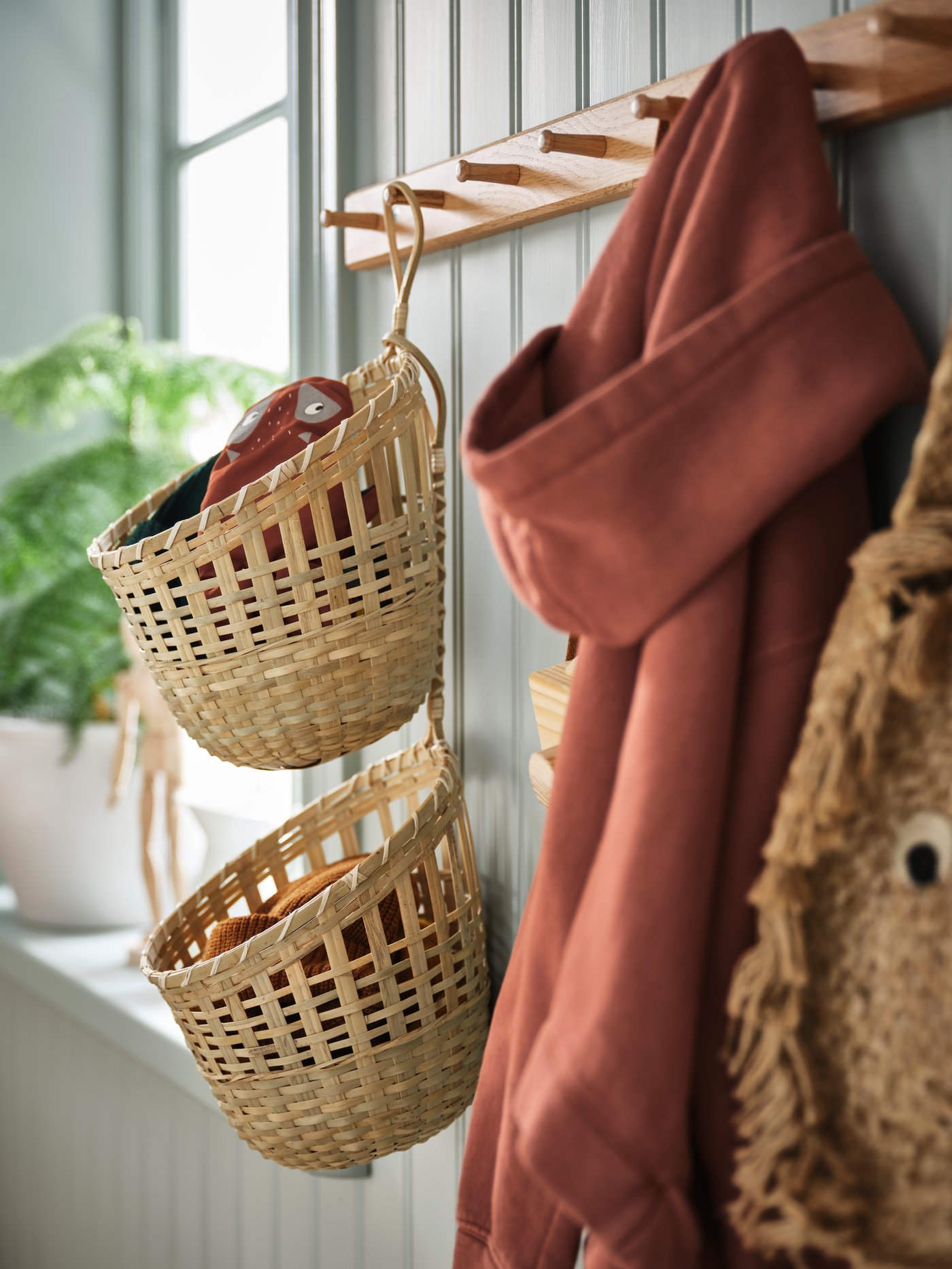 Su una parete salvia a listarelle c'è un attaccapanni in legno HÖVOLM. Ai ganci sono appesi due cestini portaoggetti TOLKNING e una felpa con cappuccio rossa.