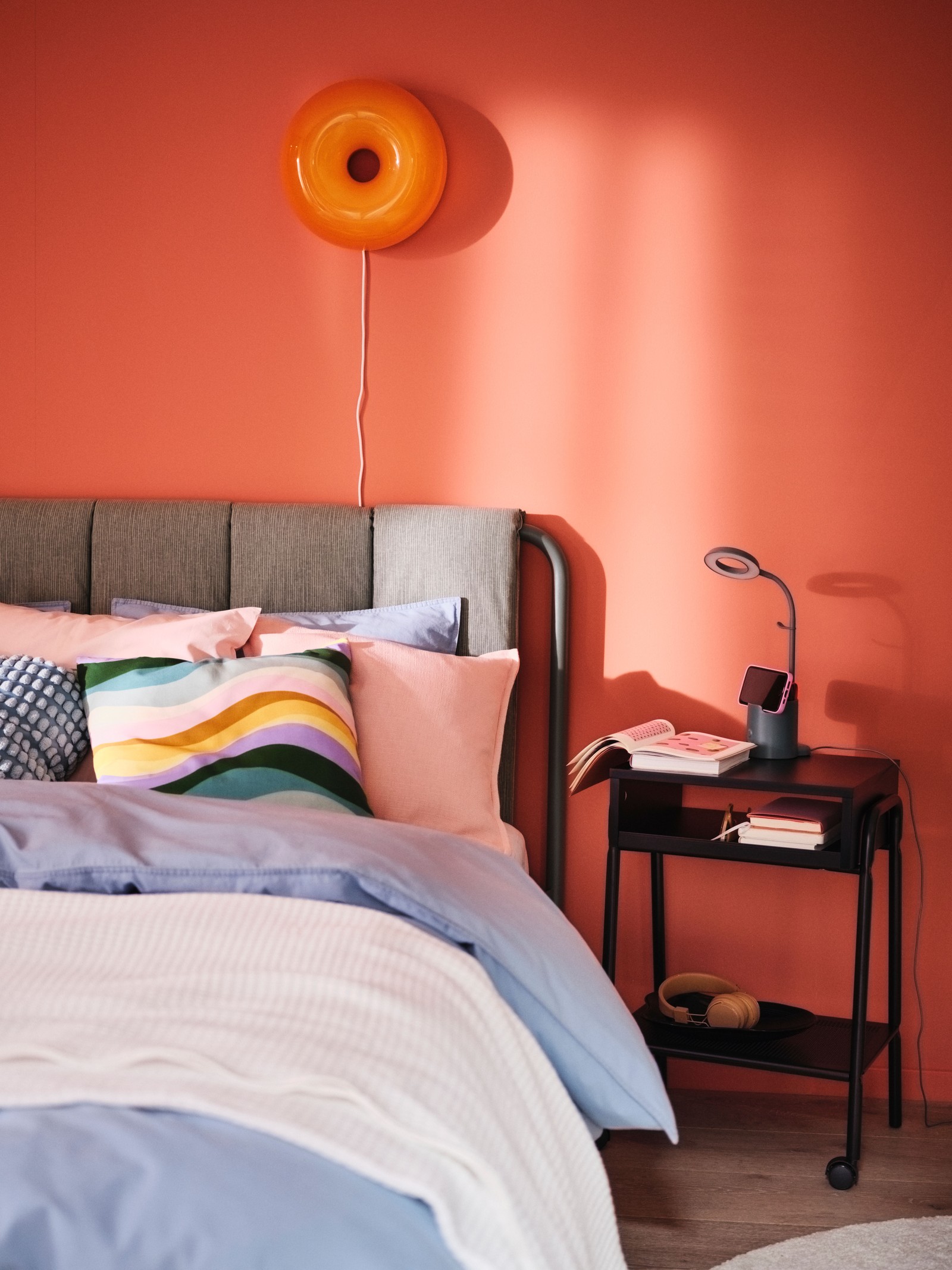 Un pat tapițat TÄLLÅSEN, aranjat cu așternuturi ÄNGSLILJA albastru-gri. Pe perete este montată o veioză/aplică LED VARMBLIXT.