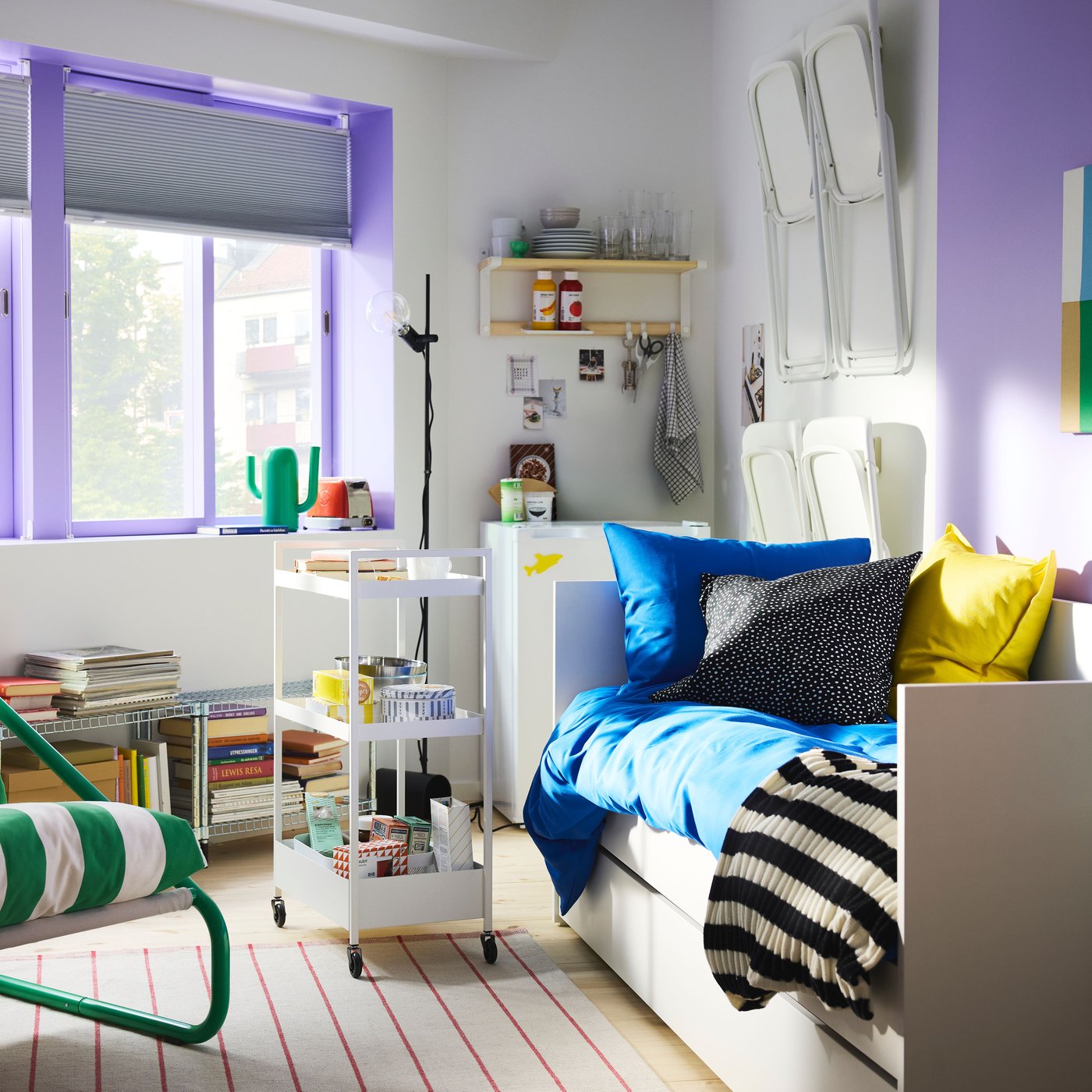 Camera da letto con grandi finestre con telaio dipinto di viola, un letto divano bianco, un carrello con rotelle e una poltrona ÖNNESTAD verde/bianca.