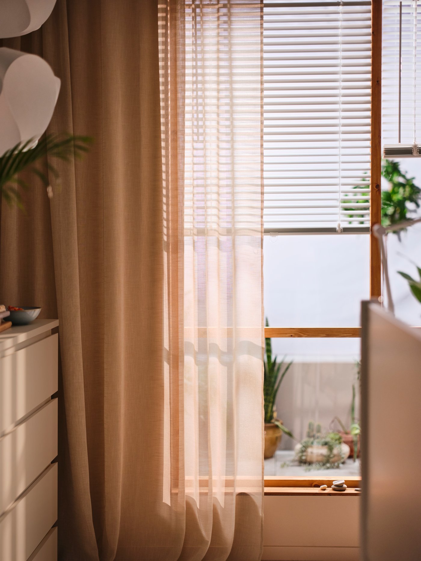 A white VECKLARFLY venetian blind half open on a window with a HÄLLEBRÄCKA sheer curtain in light beige in front of it.