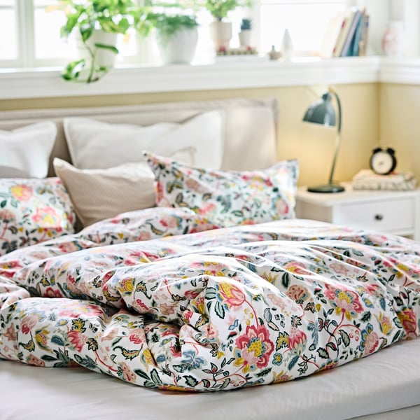 Funda nórdica DAGSSALVIA con un estampado floral extendida sobre una cama en un dormitorio, con cojines y elementos decorativos en el alféizar de la ventana.