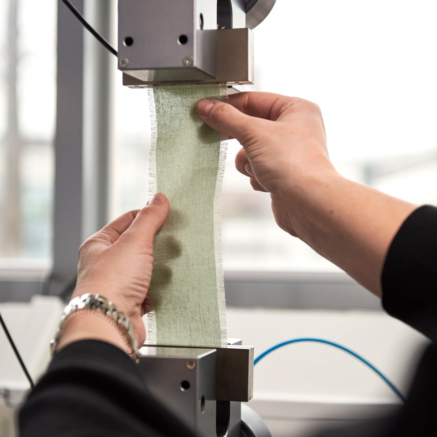 A close-up of hands arranging a cut strip of a pale grey-green ÄNGSLILJA duvet cover and pillowcase in a testing machine.