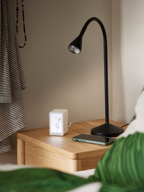 Bedroom corner with a black NÄVLINGE LED work lamp placed on a TONSTAD bedside table next to a VINDSTYRKA air quality sensor.