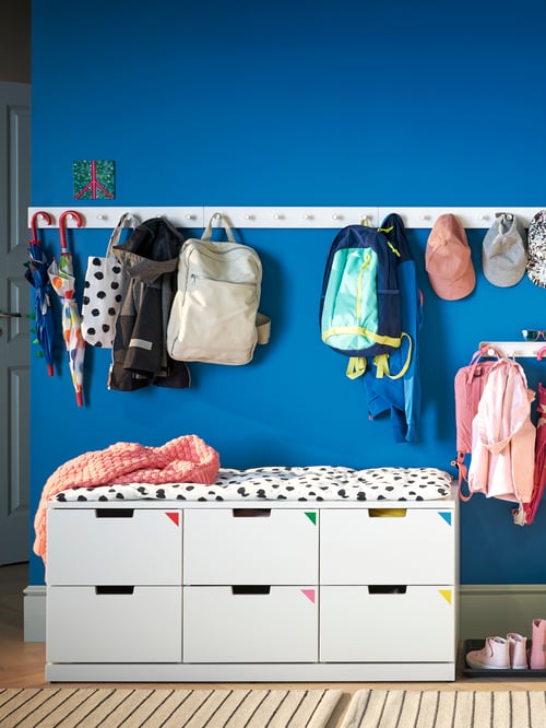 Hallway with blue wall, with six-drawer chest, multiple racks, umbrellas, backpacks, caps, and clothes.