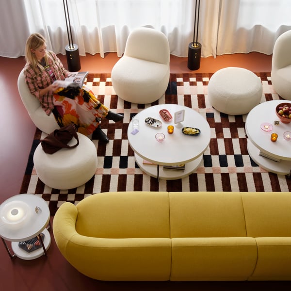A person sits across from a round, white glass FRÖTORP coffee table holding snacks in a hotel lounge with other curvy seating.