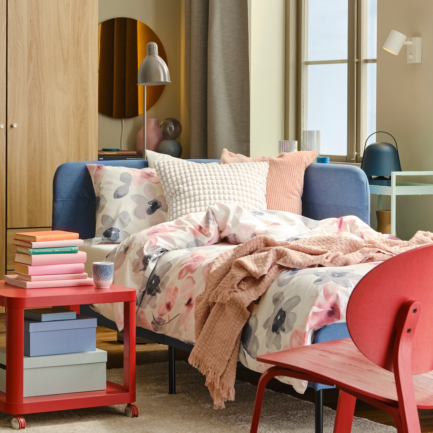 Un letto BLÅKULLEN blu con testiera angolare preparato con biancheria da letto LAPPNYCKLAR in una stanza luminosa. In fondo alla stanza un armadio con affianco una lampada da tavolo e uno specchio. Accanto al letto un tavolino con rotelle TINGBY su tappeto STOENSE. A lato un tavolo grigio con delle sedie blu.