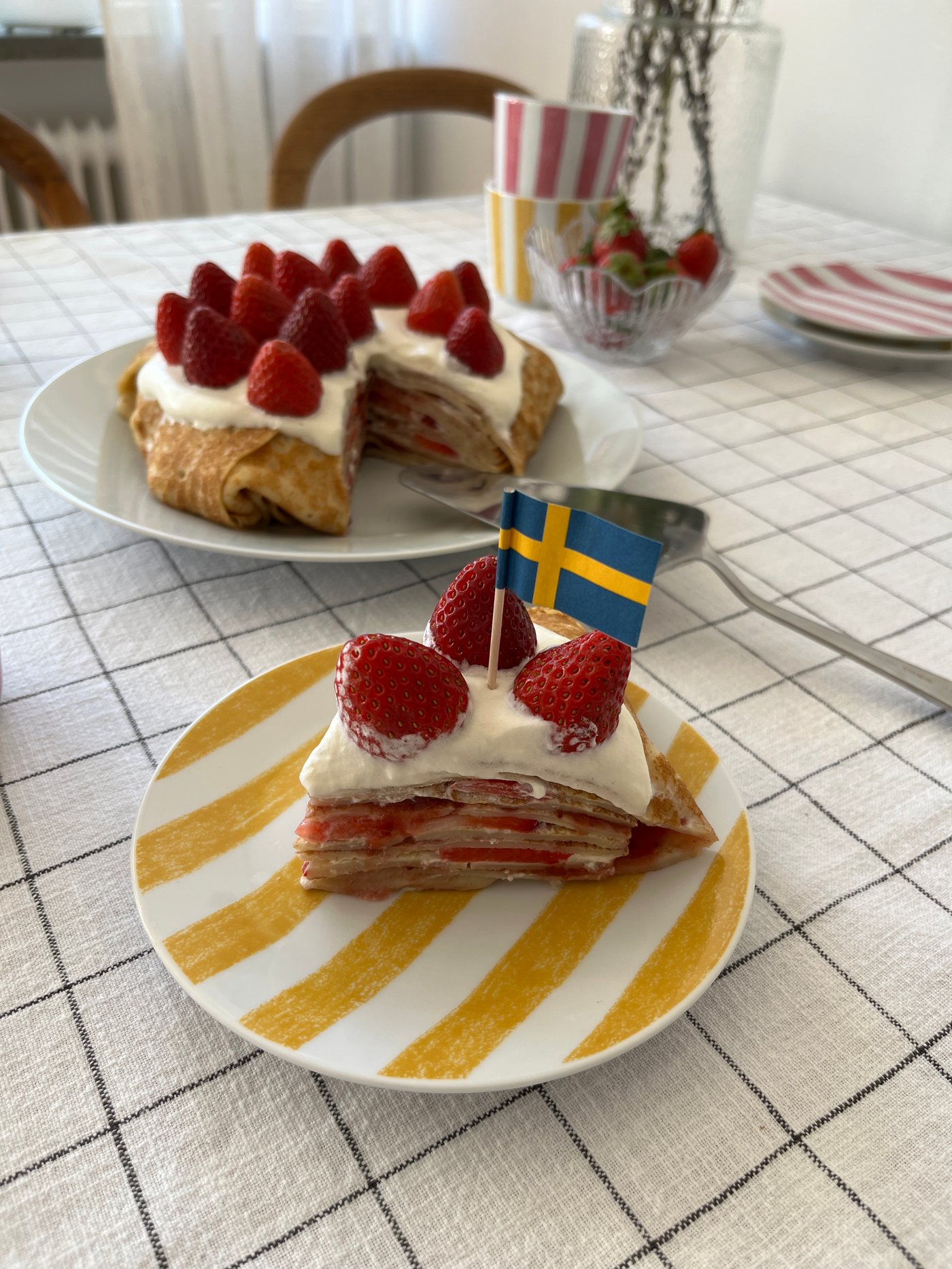 På en KLIPPAKLEJA assiett ligger en bit pannkakstårta med en Sverige-flagga i. I bakgrunden syns resten av tårtan på ett vitt fat.