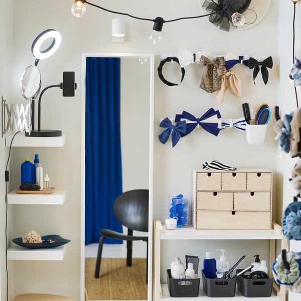 A birch plywood MOPPE mini chest on a white shelf with hair acessories on top next to a white NISSEDAL mirror on the wall.