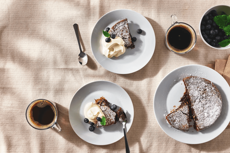 Plakken cake met poedersuiker, room en blauwe bessen, geserveerd met kopjes koffie op lichte tafel in bovenaanzicht.