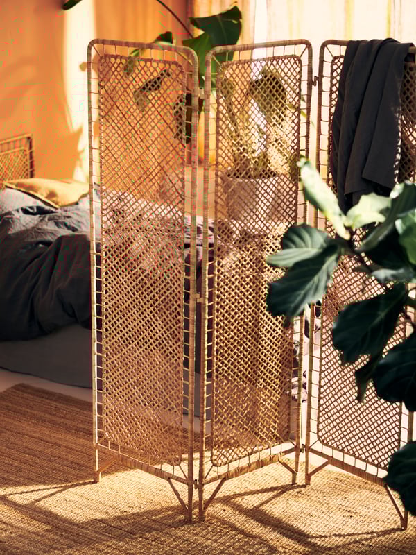 Ein stilvoller Raumteiler aus Rattan, der das Schlafzimmer harmonisch trennt. Die natürliche Textur des Rattans fügt sich perfekt in das warme, sonnendurchflutete Ambiente ein. Der Raumteiler ist praktisch und dekorativ zugleich, während er gleichzeitig Platz für Pflanzen und Kleidung bietet, die das gemütliche, einladende Gefühl des Zimmers verstärken.