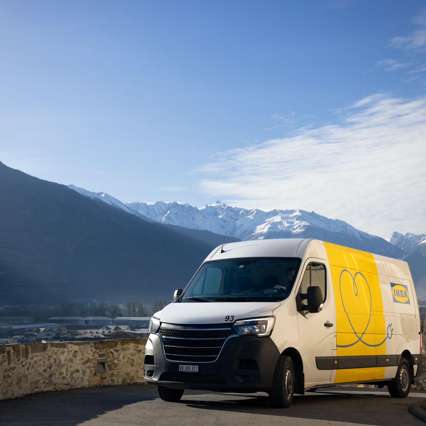 Et eldrevet køretøj med en IKEA levering på en vej i Schweiz. Himlen er blå, og der er en delvist sneklædt bjergkæde i baggrunden.