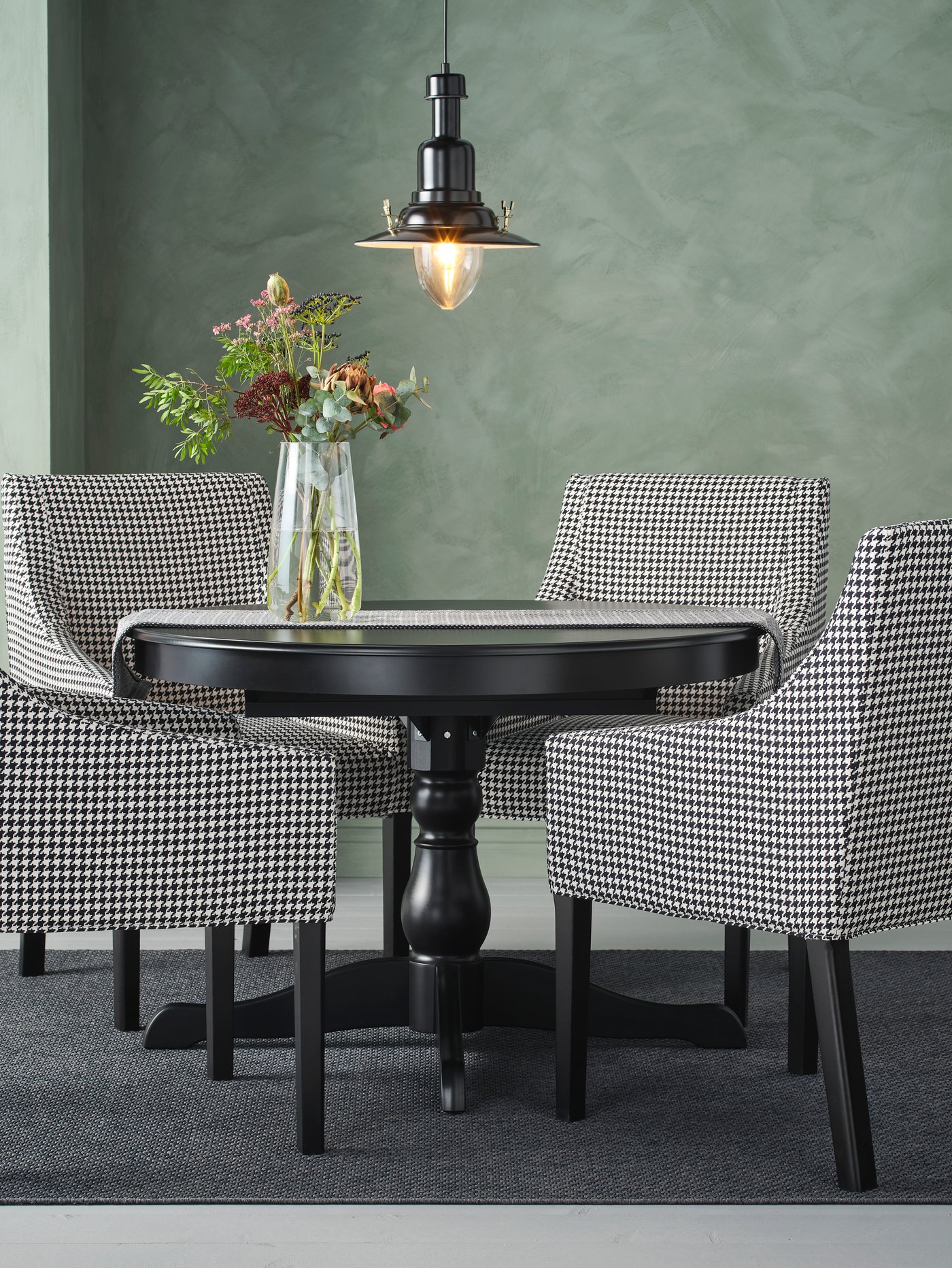 Zona de comedor con mesa de comedor extensible negra, lámpara de techo, alfombra lisa, jarrón de vidrio con flores y sillas con reposabrazos.