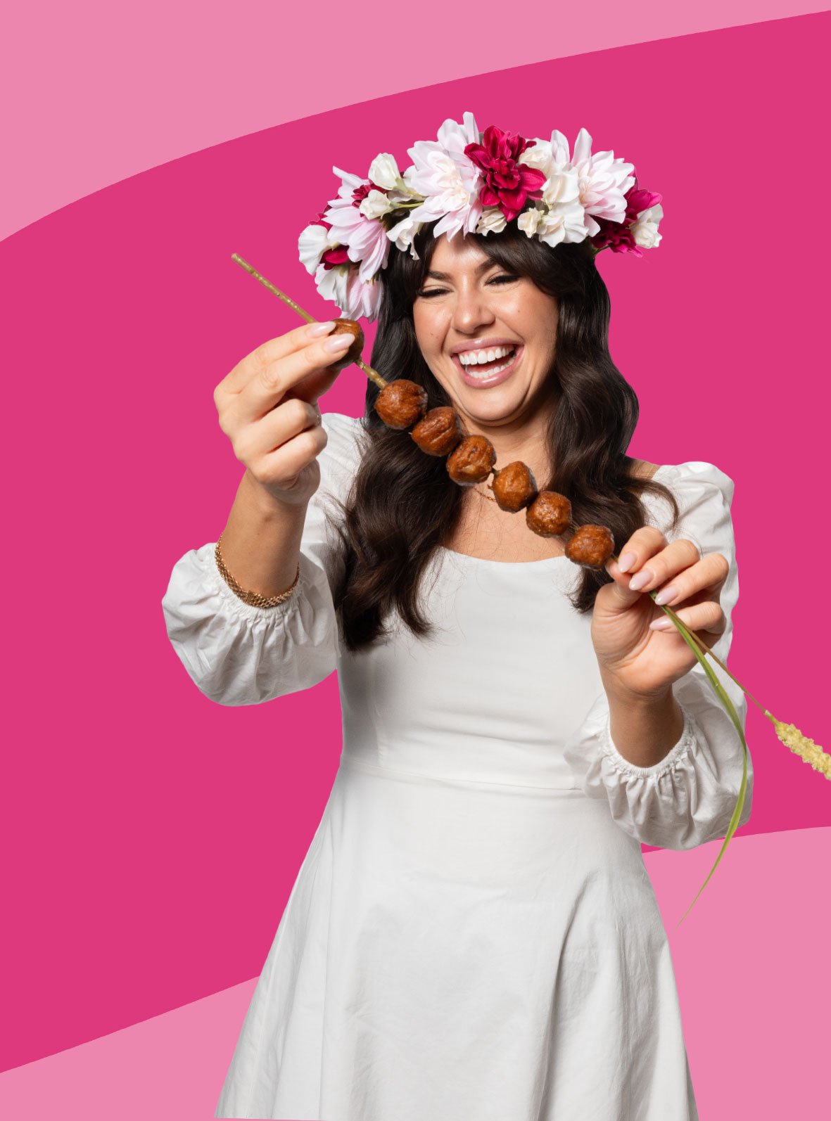 Das Bild zeigt eine Person in einem weißen Kleid mit Blumenkranz auf dem Kopf, die eine Holzspießkette mit Fleischbällchen in die Kamera hält. Der Hintergrund ist in kräftigen Pinktönen gestaltet.