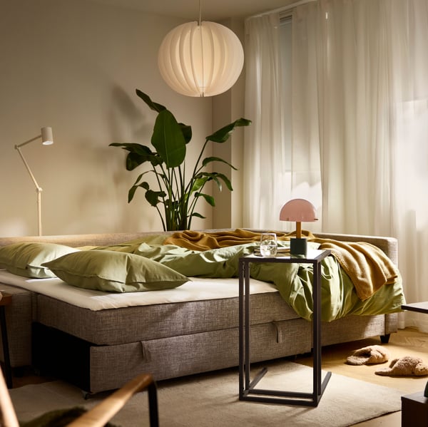 Ein Ecksofa mit Schlaffunktion von FRIHETEN/KLAGSHAMN, hier als Bett dargestellt, steht in einem sanft beleuchteten Wohnzimmer mit einem Teppich von VOLLERSLEV.