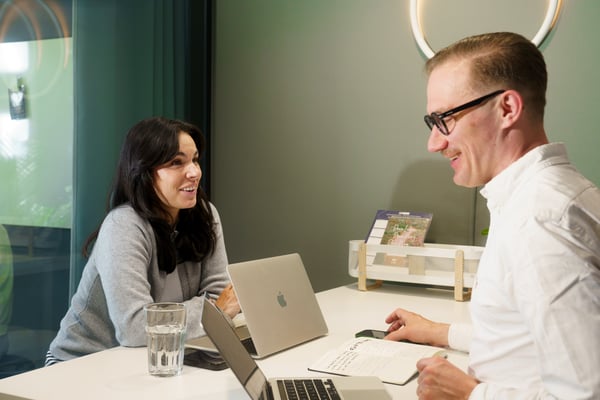 Zwei Personen sitzten in einem modern beleuchteten Besprechungsraum mit ihren Laptops auf einem Tisch zwischen ihnen.