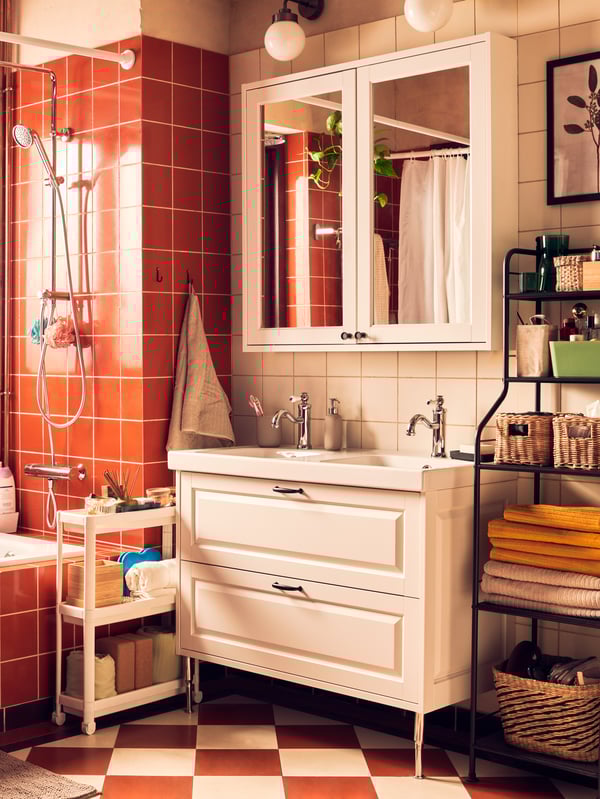 The image displays a cozy bathroom corner with terracotta tiles on the walls and a white-tiled floor. A white porcelain sink with a cabinet below is mounted against the wall, accompanied by two silver faucets. Above the sink, there’s a small open window with white frames, revealing a glimpse of greenery outside. To the left of the sink stands a shower area with an attached showerhead and hose, without any visible shower curtain or glass partition. On the right side of the sink, there’s a metal shelving unit holding various bathroom items like towels, toiletries, and containers. A beige towel hangs on the side of this unit. In front of the shelving unit is a wicker basket on the floor. The warm lighting and earthy tones give off an inviting atmosphere.