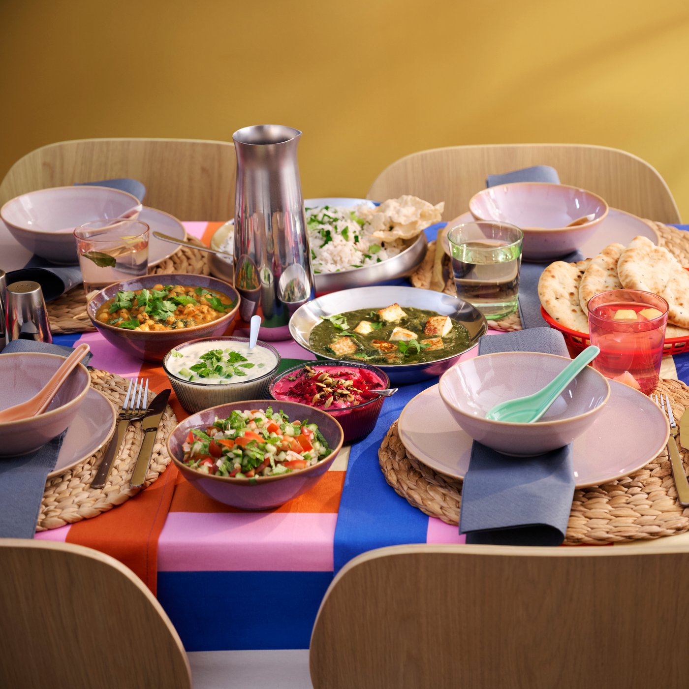 A table has a tablecloth with a colourful pattern and there are colourful bowls and plates filled with food placed on it.