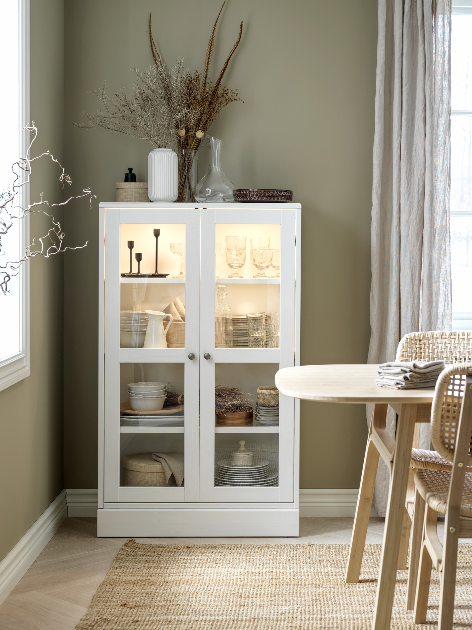 Bela vitrina sa staklenim vratima u trpezariji, drži naslagane tanjire, crne svećnjake, keramičke bokale i druge dodatke u skandinavskom deko stilu.
