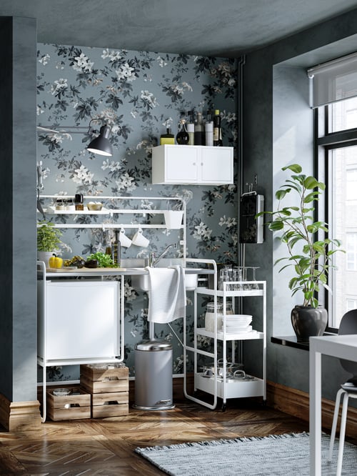 A SUNNERSTA mini-kitchen against a wall with a floral wallpaper. Vegetables and herbs are being prepared on a chopping board.