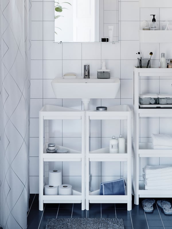 Two white VESKEN corner shelf units are placed underneath a white wash-basin and there are bathroom products on the shelves.