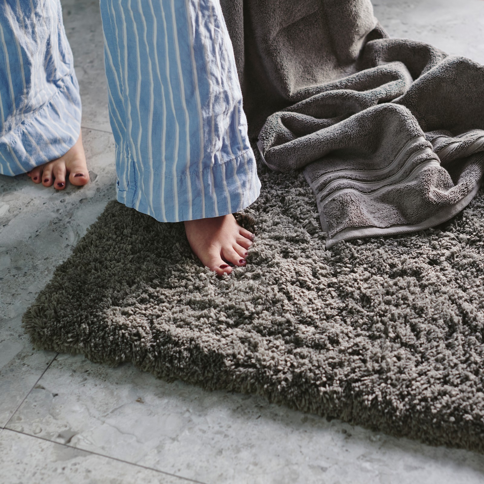 A person is stepping next to a grey bath towel onto a dark grey ALMTJÄRN bath mat on a light grey, marble-effect tiled floor.