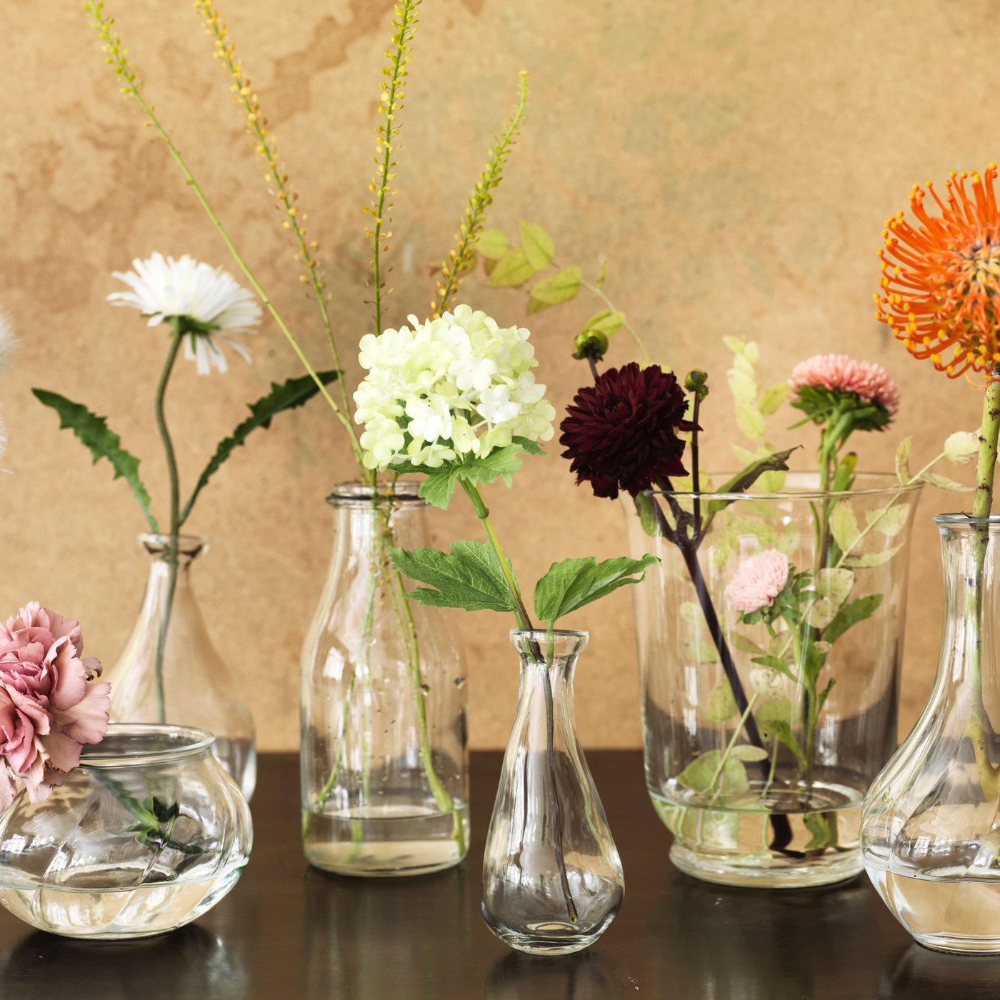 Pöydällä on useampi läpinäkyvä lasinen maljakko, joista useampaan on asetettu vain yksi näyttävä kukka.